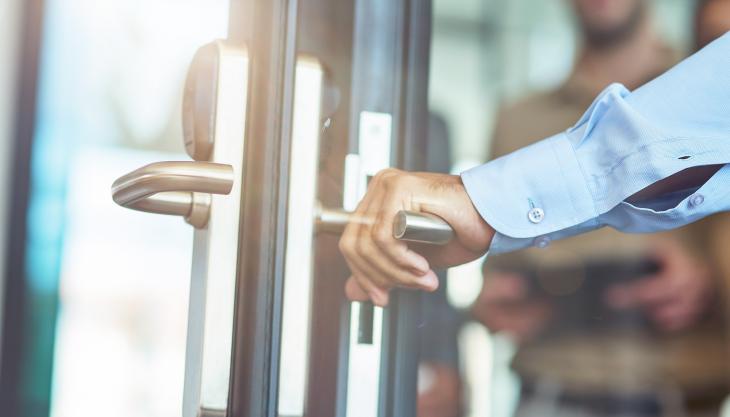 Die Hand eines Mannes im bürotauglichen Hemd umfasst die Klinke einer Bürotür.