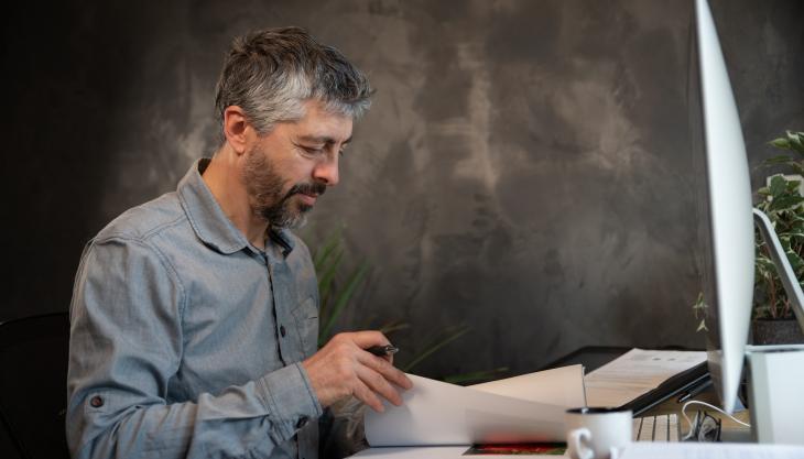 Ein Mann im grauen Businesshemd sitzt konzentriert am Schreibtisch und arbeitet sich durch Dokumente in Papierform.
