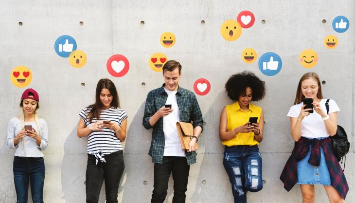 5 junge Menschen stehen entspannt vor einer Wand bzw. lehnen sich teilweise daran und schauen auf ihre Handys, aus denen Emojis aufsteigen.