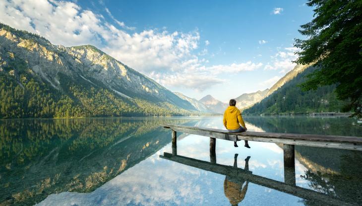 Eine Person sitzt auf dem Steg eines glasklaren Bergsees.