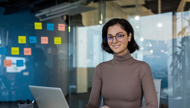 Eine junge Frau sitzt am Laptop, im Hintergrund hängen Post-its an einer Glaswand.