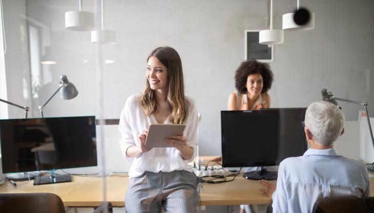 3 Mitarbeitende stehen oder sitzen in einem Büro mit Holztischen und Computerbildschirmen.