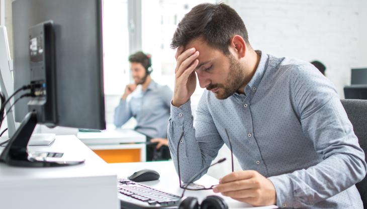 Ein Mann sitzt mit einem anderen in einem Büro am Tisch und stützt müde den Kopf in die Hand.