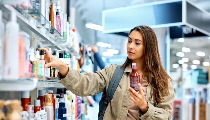 Eine junge Frau in einer Drogerie, sie nimmt eine Tube aus dem Regal und hält eine andere in der Hand.