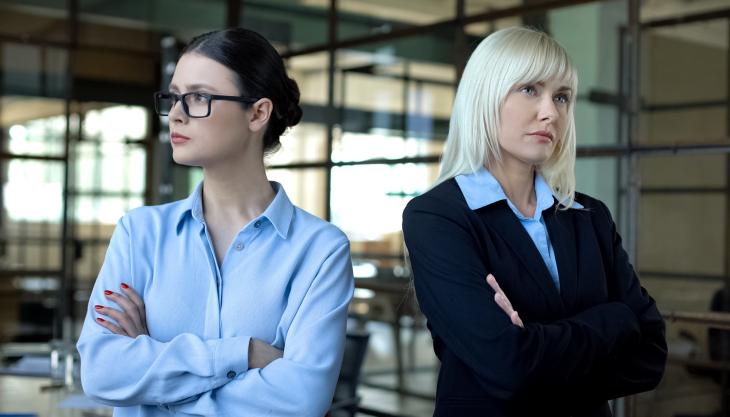 Zwei Frauen in Businesskleidung stehen mit ernsten Gesichtern und verschränkten Armen einander abgewandt.