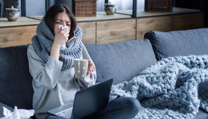 Eine schwarzhaarige Frau sitzt vor einem Laptop auf dem Sofa und putzt sich die Nase, in der Hand hat sie eine Teetasse.