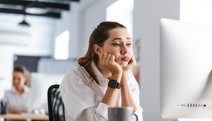 Eine junge Frau sitzt gelangweilt vor einem weißen PC-Bildschirm, im Hintergrund sitzt eine andere Mitarbeiterin.