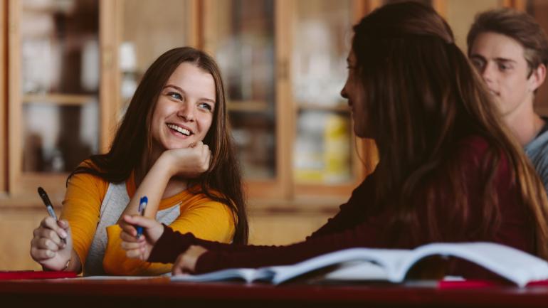 Zwei Studentinnen und ein Student sitzen lächelnd zusammen und lernen.