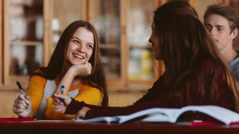 Foto: Canva.com Zwei Studentinnen und ein Student sitzen lächelnd zusammen und lernen.