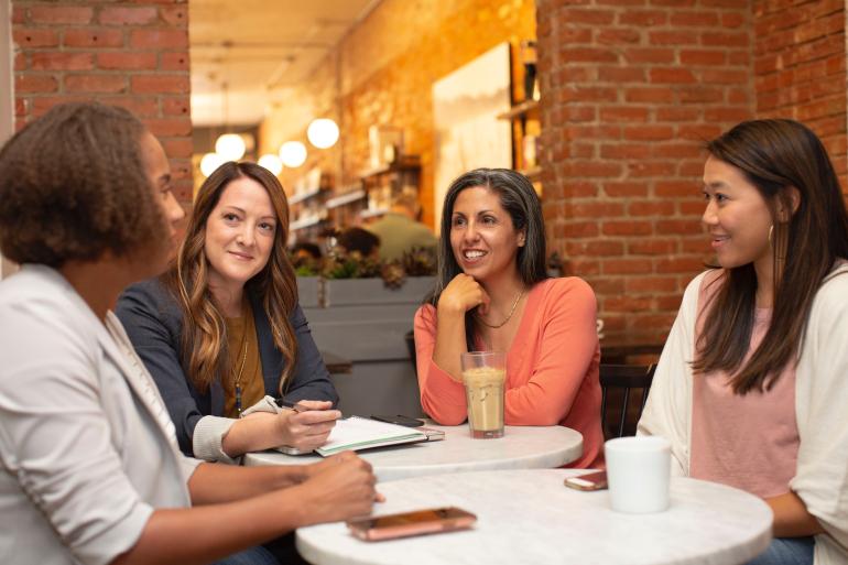 Vier Frauen sitzen an einem Tisch und und unterhalten sich lächelnd.