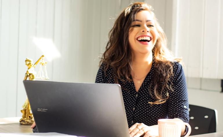 Brünette junge Frau mit Locken sitzt lachend vor ihrem Laptop.