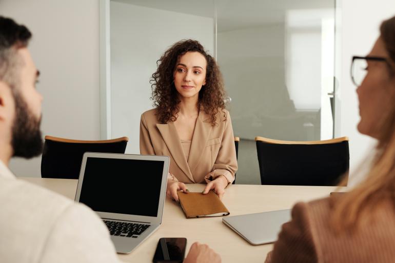 Auch durch nonverbale Signale beeinflussen Sie die Verhandlungssituation (Foto: Edmond Dantès / Pexels.com). Eine Frau sitzt am Tisch gegenüber eines Mannes und einer anderen Frau. Auf dem Tisch steht ein Laptop.