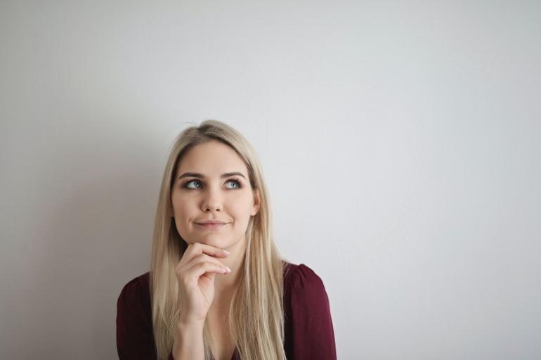 Eine Frau sitzt vor einer weißen Wand. Sie hat eine Hand am Kinn in nachdenklicher Stellung.