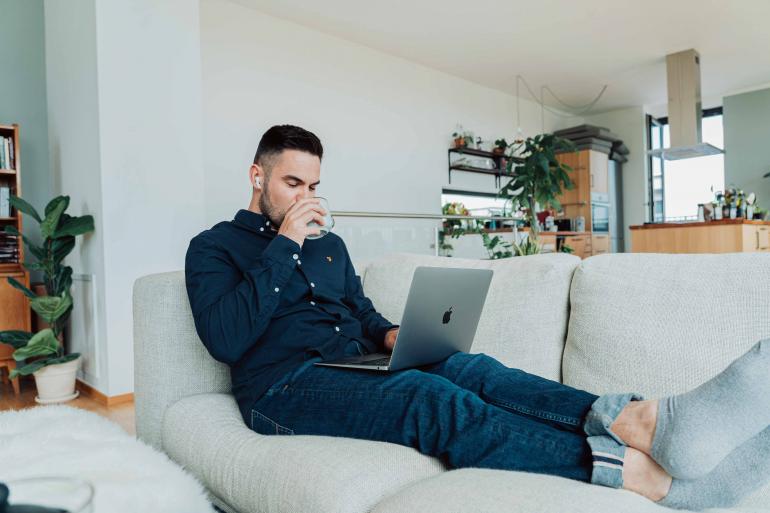 Mann sitzt auf Sofa mit Laptop auf einen Knien. ER trink aus einer Tasse. 