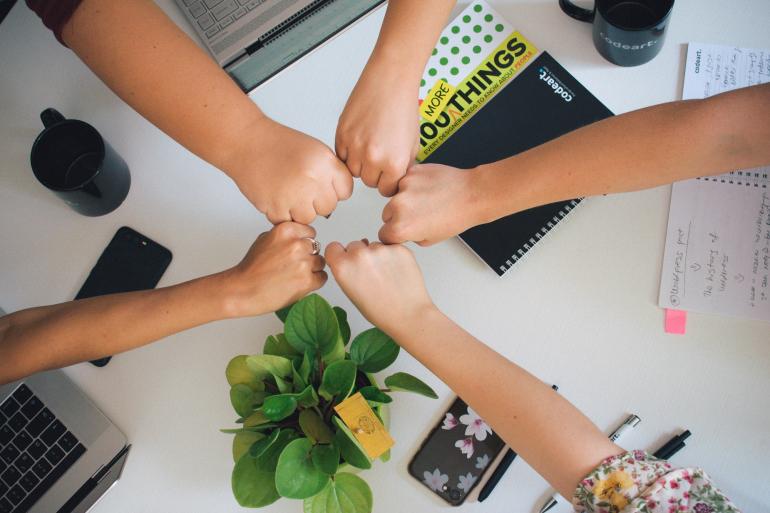 Foto: Antonio Janeski - Unsplash.com Über einem Schreibtisch mit Notebooks und Notizheften strecken vier Menschen ihre rechte Hand aus, die zur Faust geballt ist, und berühren sich mit ihren Fäusten in der Mitte des Bildes.