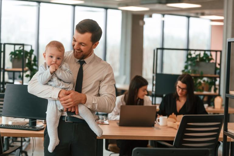 Ein junger Vater im Business-Anzug steht im Büro und hält lachend sein Baby auf dem Arm.