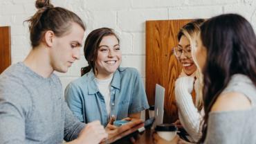 Drei junge Frauen und ein junger Mann sitzen lächelnd zusammen am Tisch und schauen auf ein Tablet.