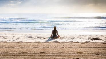 Frau sitzt im Schneidersitz am Strand mit Blick auf das Meer und meditiert.