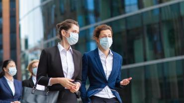 Zwei Frauen im Business-Outfit laufen mit Maske nebeneinander her.