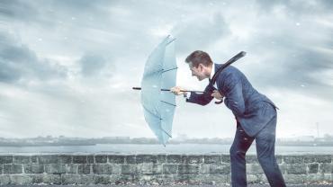 Ein Mann im Business-Anzug steht bei stürmischem Wetter draußen, hält seinen aufgespannten Schirm vor sich und beugt sich damit gegen Wind und Regen.