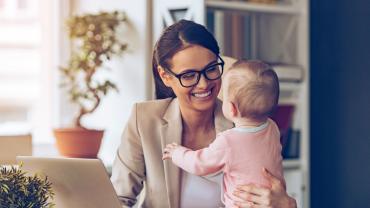 Eine Mutter im Businessoutfit sitzt am Laptop und lächelt ihr Baby an, das neben ihr auf der Tischplatte sitzt und das sie mit dem linken Arm festhält.