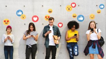 5 junge Menschen stehen entspannt vor einer Wand bzw. lehnen sich teilweise daran und schauen auf ihre Handys, aus denen Emojis aufsteigen.