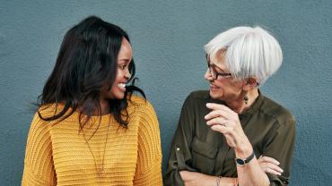 Eine junge schwarze Frau und eine ältere weiße Frau stehen im lockeren Business-Outfit nebeneinander und lachen sich an.