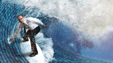 Fotomontage: Ein Mann im Businessoutfit steht auf einem Surfbrett und surft auf einer Datenwelle.
