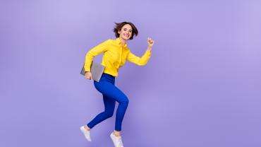 Eine junge Frau im sportlichen Business-Outfit springt mit dem Laptop in der Hand nach vorn oben in die Luft.