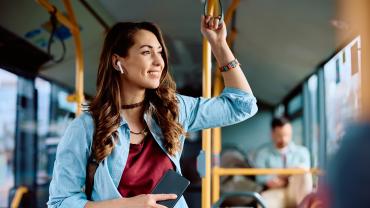 Eine junge Frau steht im Bus und hört über Bluetooth-Ohrstöpsel einen Podcast.