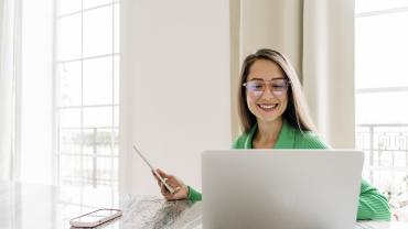 Eine junge Frau sitzt an einem Tisch vor ihrem Laptop und hält ein Tablet in der Hand. Auf dem Tisch liegt ein Smartphone.