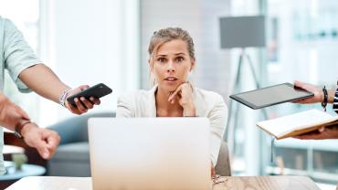 Eine junge Frau sitzt im Office am Laptop und von allen Seiten werden ihr Aufgaben in Form von Tablets, Handys, Papieren gereicht.