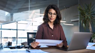 Eine Frau mit dunklen Haaren sitzt im Büro an einem Laptop und schaut auf ein Blatt Papier.