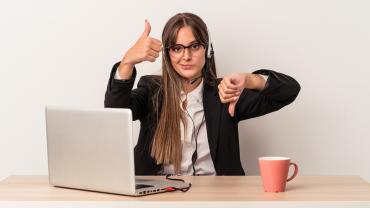 Eine junge Frau sitzt an einem Laptop und streckt einen Daumen nach oben und einen nach unten.