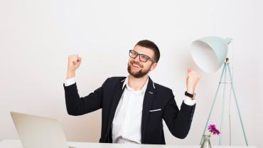 Ein junger Mann im Anzug sitzt an einem Büroschreibtisch und ballt triumphierend die Hände.