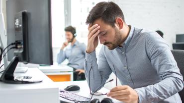 Ein Mann sitzt mit einem anderen in einem Büro am Tisch und stützt müde den Kopf in die Hand.