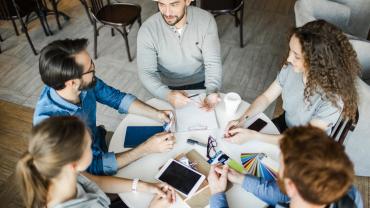 5 Mitarbeitende sitzen an einem runden Tisch mit Arbeitsmaterialien