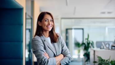 Eine junge Frau in Blazer steht in einem Büro und lächelt selbstbewusst in die Kamera.