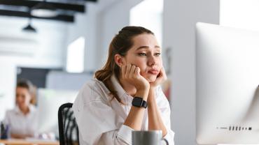 Eine junge Frau sitzt gelangweilt vor einem weißen PC-Bildschirm, im Hintergrund sitzt eine andere Mitarbeiterin.