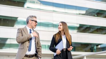 Ein mittelalter Mann und eine junge Frau in Businesskleidung laufen nebeneinander, im Hintergrund sieht man ein Bürogebäude.