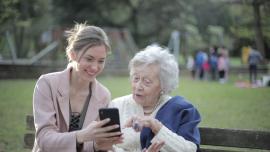 Junge und ältere Frau sitzen zusammen auf einer Parkbank und die junge Frau zeigt der älteren etwas auf einem Smartphone.