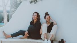 Zwei Frauen sitzen lachend nebeneinander auf dem Boden, an eine weiße Wand gelehnt.