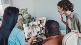 Drei Mitarbeitende stehen bzw. sitzen um einen Bildschirm, auf dem eine Videokonferenz zu sehen ist.