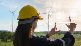 Eine Ingenieurin mit Bauhelm steht auf einem grünen Feld, im Hintergrund sind Windräder zu sehen. Sie hält ihre Hände vor sich und blickt auf einen transparenten Bildschirm mit diversen Buttons, den sie vor ihrem geistigen Auge sieht.