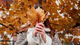 Eine junge Frau im Mantel hält sich Herbstblätter vor das Gesicht, im Hintergrund steht ein Baum mit Herbstlaub