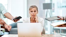 Eine junge Frau sitzt im Office am Laptop und von allen Seiten werden ihr Aufgaben in Form von Tablets, Handys, Papieren gereicht.