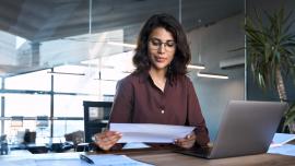 Eine Frau mit dunklen Haaren sitzt im Büro an einem Laptop und schaut auf ein Blatt Papier.
