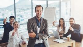 4 Mitarbeitende sitzen an einem Tisch und recken den Daumen nach oben, am Tisch lehnt ein Manager und hat die Arme lächelnd verschränkt.