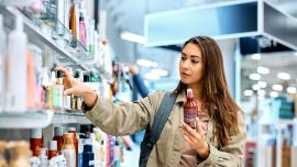 Eine junge Frau in einer Drogerie, sie nimmt eine Tube aus dem Regal und hält eine andere in der Hand.