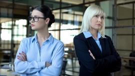 Zwei Frauen in Businesskleidung stehen mit ernsten Gesichtern und verschränkten Armen einander abgewandt.
